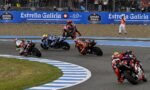 MotoGP-style race in progress: riders lean into a sharp turn on a blue-striped track, Estrella Galicia banners and a cheering crowd in the background.