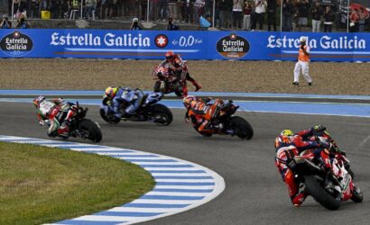 MotoGP-style race in progress: riders lean into a sharp turn on a blue-striped track, Estrella Galicia banners and a cheering crowd in the background.