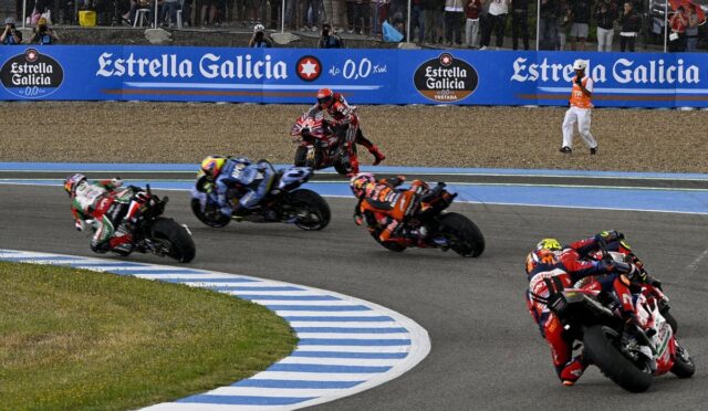 MotoGP-style race in progress: riders lean into a sharp turn on a blue-striped track, Estrella Galicia banners and a cheering crowd in the background.