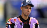 MotoGP rider in a purple racing suit and navy cap stands on a track, looking to the side.