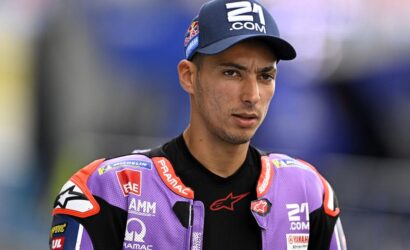 MotoGP rider in a purple racing suit and navy cap stands on a track, looking to the side.