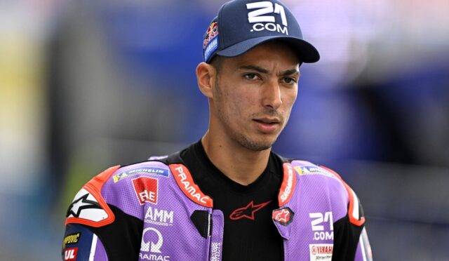 MotoGP rider in a purple racing suit and navy cap stands on a track, looking to the side.
