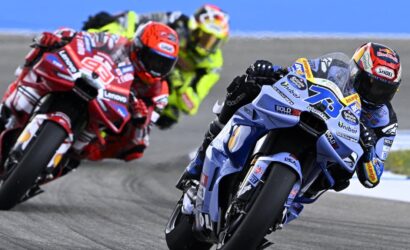Blue and white race bike #75 leads a pack of riders leaning into a turn on a racetrack, helmeted and focused on the curve.