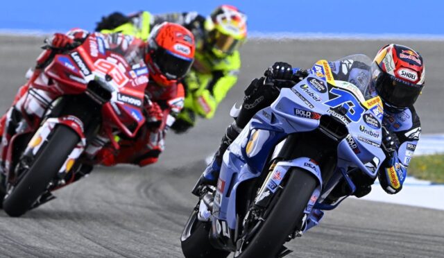 Blue and white race bike #75 leads a pack of riders leaning into a turn on a racetrack, helmeted and focused on the curve.