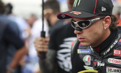 Moto racer in black cap and white sunglasses, focusing ahead, in a paddock with blurred crowd behind him.