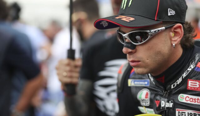 Moto racer in black cap and white sunglasses, focusing ahead, in a paddock with blurred crowd behind him.