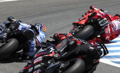 Group of motorcycle racers leaning into a sharp turn on a race track, blue and red suits, blue-and-white curbing visible.