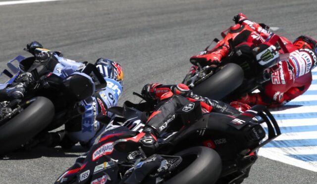 Group of motorcycle racers leaning into a sharp turn on a race track, blue and red suits, blue-and-white curbing visible.