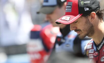 MotoGP rider in a red cap with sponsor logos looking downward, wearing a red racing suit with patches like Lenovo and Monster Energy