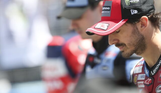 MotoGP rider in a red cap with sponsor logos looking downward, wearing a red racing suit with patches like Lenovo and Monster Energy
