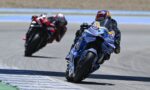 Two motorcycle racers lean into a sharp turn on a racetrack, blue bike in the foreground.