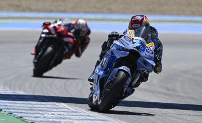 Two motorcycle racers lean into a sharp turn on a racetrack, blue bike in the foreground.