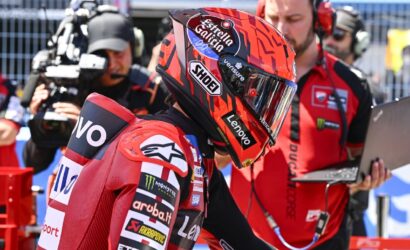 Motorcycle racer in red racing suit and helmet with sponsor logos, standing in the pit lane as a camera crew films nearby.