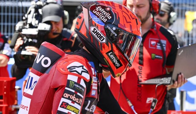 Motorcycle racer in red racing suit and helmet with sponsor logos, standing in the pit lane as a camera crew films nearby.