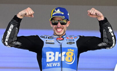Cyclist in blue race kit and cap raises fists in victory on a stage, wearing sunglasses and smiling at the crowd.