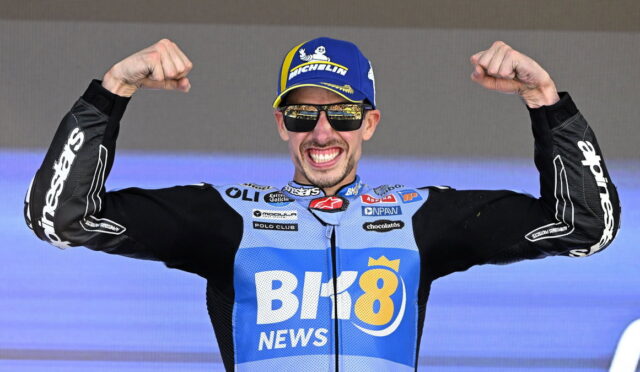 Cyclist in blue race kit and cap raises fists in victory on a stage, wearing sunglasses and smiling at the crowd.