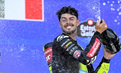 Moto racer on a podium celebrating by spraying champagne, smiling at the camera.