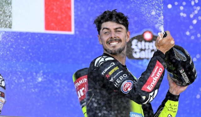 Moto racer on a podium celebrating by spraying champagne, smiling at the camera.