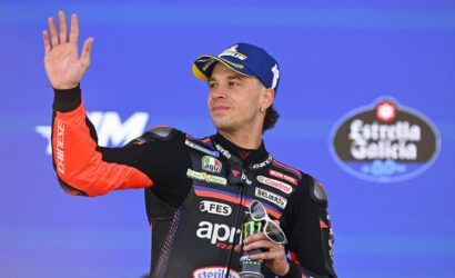 Motorcycle racer on a podium waves to the crowd, wearing an orange‑black racing suit and blue cap, holding sunglasses and a trophy