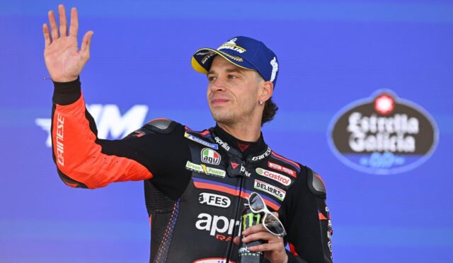 Motorcycle racer on a podium waves to the crowd, wearing an orange‑black racing suit and blue cap, holding sunglasses and a trophy
