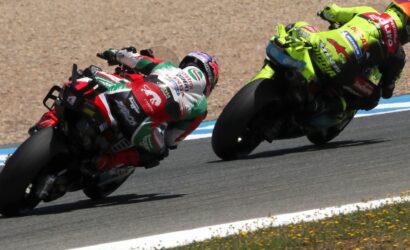 Two motorcycle racers in full leathers lean into a curve on a racetrack, one in red‑green‑white and the other in neon yellow, with a gravel runoff nearby.