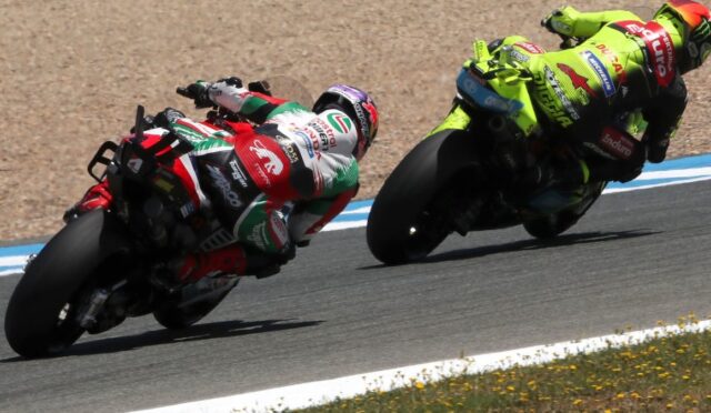 Two motorcycle racers in full leathers lean into a curve on a racetrack, one in red‑green‑white and the other in neon yellow, with a gravel runoff nearby.