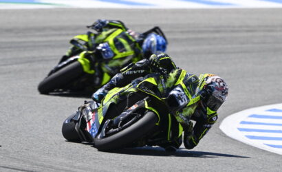Two motorcycle racers in neon yellow-and-black gear lean into a sharp track corner, one ahead with a blue sky behind.