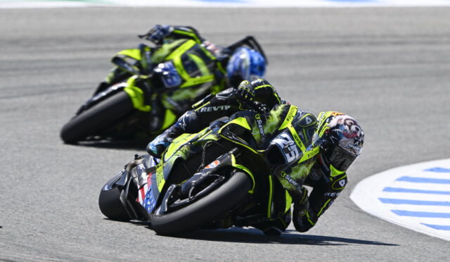 Two motorcycle racers in neon yellow-and-black gear lean into a sharp track corner, one ahead with a blue sky behind.