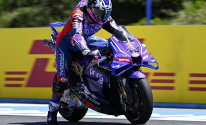 Rider in purple racing suit on a purple motorcycle leaning into a turn on a track, number 43 visible on the bike with yellow barriers in the background.