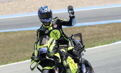 Racer on a neon-yellow and black sport motorcycle waves to the camera on a race track.