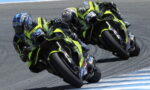 Three neon-yellow racing motorcycles lean into a sharp left turn on a race track, riders in matching leathers closely grouped.
