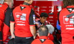 MotoGP rider in red racing suit sits in a team pit area, surrounded by crew members in matching red uniforms.