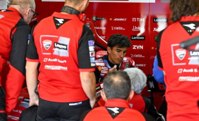 MotoGP rider in red racing suit sits in a team pit area, surrounded by crew members in matching red uniforms.