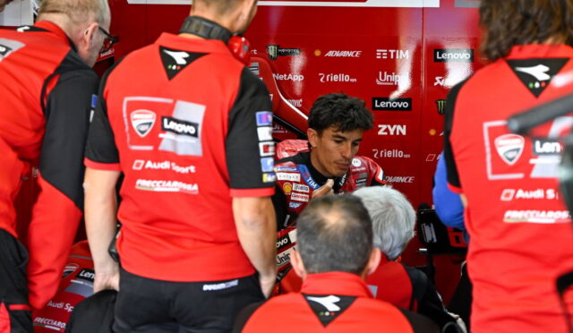 MotoGP rider in red racing suit sits in a team pit area, surrounded by crew members in matching red uniforms.