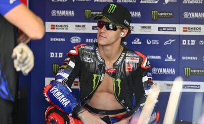 MotoGP rider in a black Monster Yamaha racing suit and cap sits in a paddock with sponsor boards behind him.