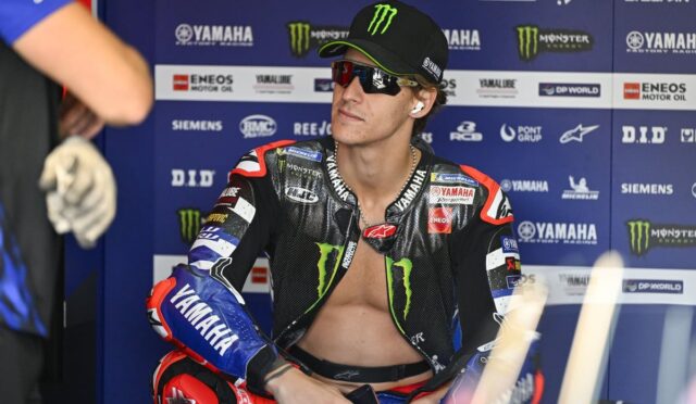 MotoGP rider in a black Monster Yamaha racing suit and cap sits in a paddock with sponsor boards behind him.