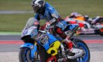 Moto racer in blue leathers leaning into a turn on a blue Honda sportbike adorned with Estrella Galicia and Bridgestone logos; blurred racers in the background.