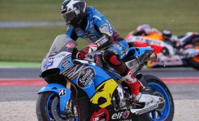 Moto racer in blue leathers leaning into a turn on a blue Honda sportbike adorned with Estrella Galicia and Bridgestone logos; blurred racers in the background.