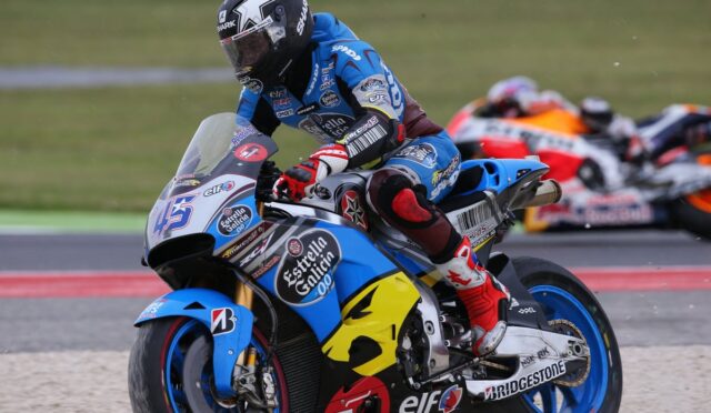 Moto racer in blue leathers leaning into a turn on a blue Honda sportbike adorned with Estrella Galicia and Bridgestone logos; blurred racers in the background.