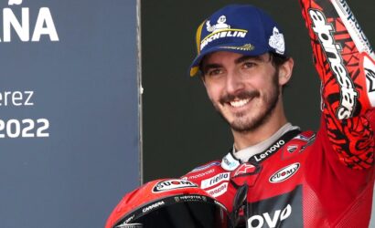 Smiling motorcycle racer in a red racing suit with a blue Michelin cap raises his arm, holding a red helmet; Jerez 2022 sign to the left.