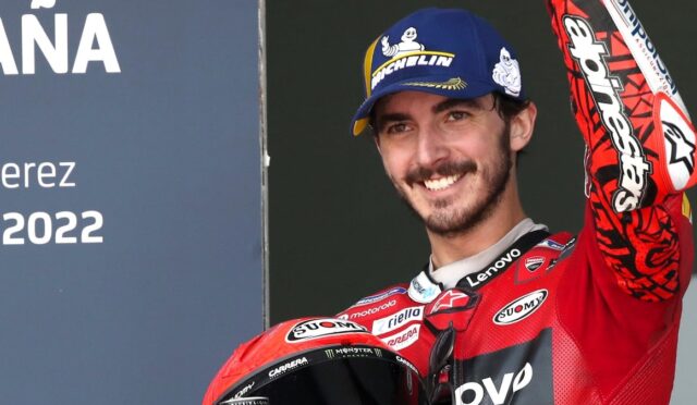 Smiling motorcycle racer in a red racing suit with a blue Michelin cap raises his arm, holding a red helmet; Jerez 2022 sign to the left.