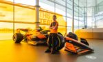 Young boy in an orange polo stands with a Formula 1 race car inside a glass-walled showroom.