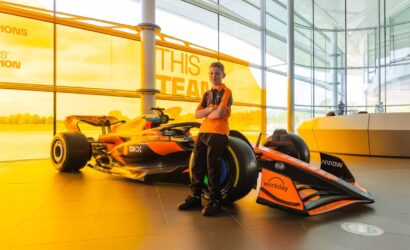 Young boy in an orange polo stands with a Formula 1 race car inside a glass-walled showroom.