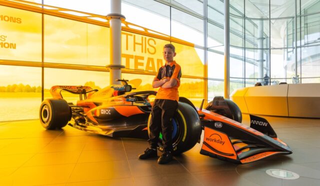 Young boy in an orange polo stands with a Formula 1 race car inside a glass-walled showroom.