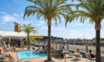 Outdoor poolside deck with palm trees, sun loungers, and guests relaxing near a bar labeled 'The Pop-Up Hotel' under a bright blue sky