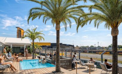 Outdoor poolside deck with palm trees, sun loungers, and guests relaxing near a bar labeled 'The Pop-Up Hotel' under a bright blue sky