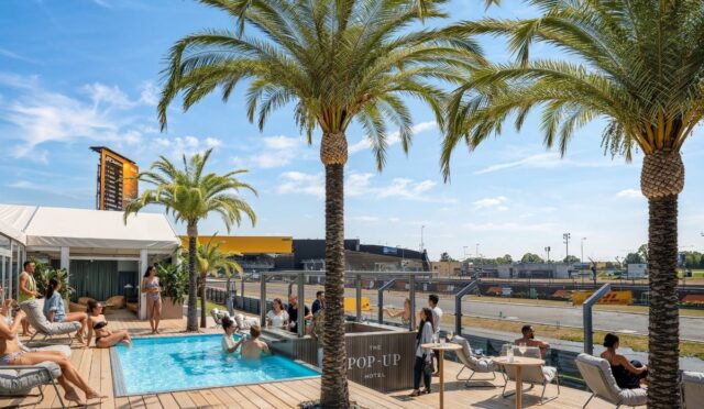 Outdoor poolside deck with palm trees, sun loungers, and guests relaxing near a bar labeled 'The Pop-Up Hotel' under a bright blue sky