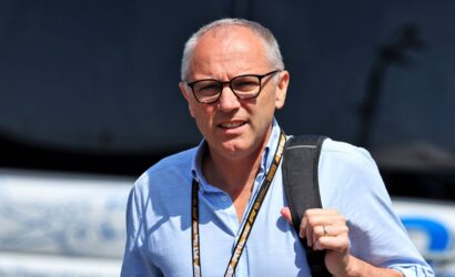 Mid-aged man with glasses in a light blue shirt, carrying a black backpack and wearing a lanyard outdoors.
