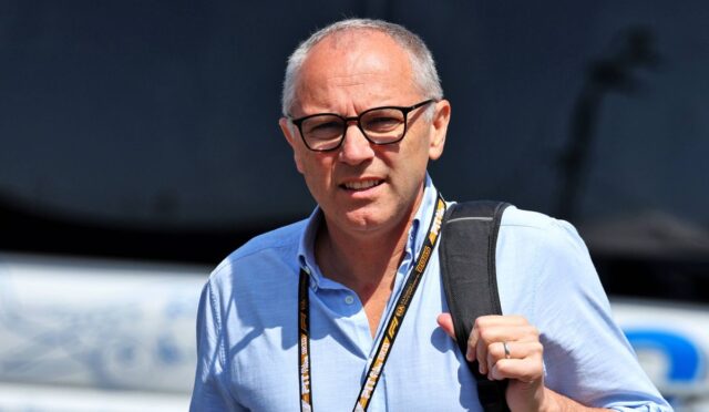 Mid-aged man with glasses in a light blue shirt, carrying a black backpack and wearing a lanyard outdoors.