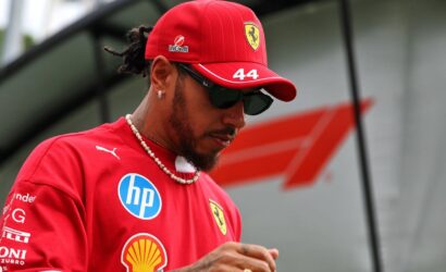 Ferrari driver wearing a red cap with number 44 and sunglasses, looking down with a beaded necklace on a red team shirt.